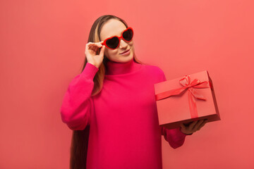 Happy smiling stylish young woman in trendy heart shaped glasses and pink knitted dress holding present gift with ribbon bow isolated on colored pink background.

Happy holiday celebration concept.