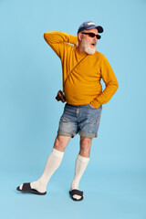 Portrait of senior bearded man in casual clothes with cap, sunglasses and vintage camera posing over blue background. Leisure