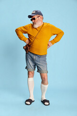 Portrait of senior bearded man in casual clothes with cap, sunglasses and vintage camera posing over blue background.