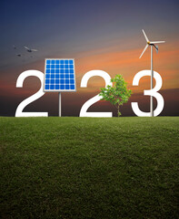 2023 white text with solar cell, wind turbine and growing tree on green grass field over sunset...