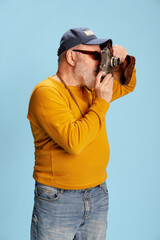 Portrait of senior bearded man in casual clothes with cap, sunglasses, taking photo with vintage camera posing over blue background. Tourist