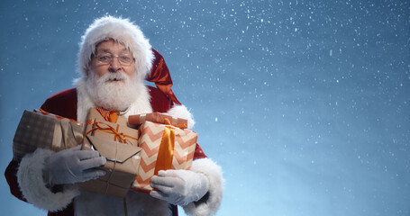 Close up shot of senior bearded man in santa clause outfit carrying gift boxes, isolated on blue...