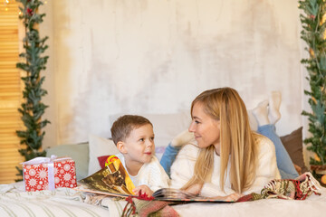 Happy family mother and son reading book together on bed on Christmas Eve. Concept of holiday joy and fun. New Year morning. Winter holidays at home