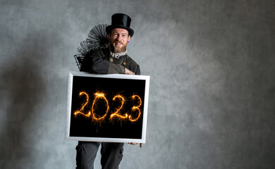Schornsteinfeger in Arbeitskleidung und mit Kehrgeräten vor grauer Wand, hält Bilderrahmen mit Jahreszahl 2023 mit Wunderkerzen geschrieben, in der Hand.