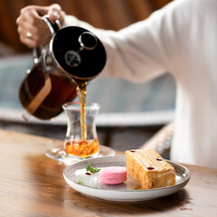 Piece of honey cake and macaron on plate with cup of tea on wooden table. Pouring tea into a cup. Turkish tea. Cutaway honey cake. Serving dish.