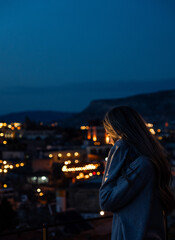 Blonde woman with hoodie looking down the cappadocia town with lights of the street in the night