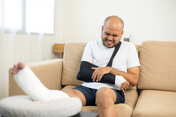 Man suffered pain from accident fracture broken bone injury with leg splints in cast neck splints collar arm splints sling support arm in living room. Social security and health insurance concept. - obrazy, fototapety, plakaty