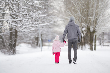 Little daughter and young adult father walking on white snow covered sidewalk at park. Spending time together in beautiful cold winter day. Enjoying stroll. Back view. - Powered by Adobe