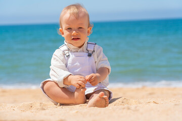 Little cheerful boy child sits on the sand on the beach. Sunny day.
