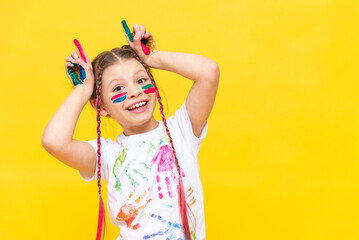 A dabbling girl stained in paints shows horns on a yellow isolated background. The child is playing with colorful paints. Courses for the development of children's creativity. Happy childhood games.
