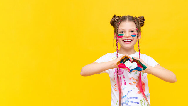 A little girl smeared with paint shows a heart. The art of drawing with multicolored paints. A child on a yellow isolated background. Copy space.