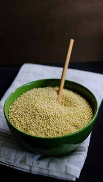 Mijo en un bol verde con una cuchara de madera dentro, encima de un trapo de cocina marr&oacute;n y blanco. Dark food