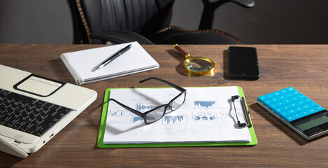 Laptop, smartphone, eyeglasses, pen, notepad, calculator, magnifying glass with a financial graphs.