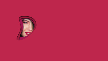 Face of young beautiful girl with a bright make-up and red lips looks through a hole in Viva Magenta paper. Closeup beauty portrait With place for text on the right
