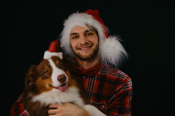 Dog and human with Santa Claus hats on their heads. Studio shot. Concept of pet celebrating Merry Christmas with male owner. Young Caucasian man with beard hugs Australian Shepherd.