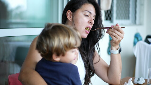 Mother Eating While Holding Child In Arms. Mom Feeding Small Boy With Fork