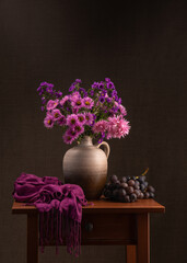 A large bouquet of pink flowers, a dark pink shawl next to it, a large bunch of blue grapes. Wooden table, gray background. Still life. Calmness, joy, good mood. Health.