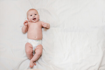 A smiling baby lies on a white bed. A small child in a diaper rejoices at the arrival of his mother and smiles.