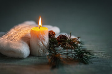 Obraz na płótnie Canvas Flame of a white candle with a knitted pattern and white knitted gloves and a pine branch with cones on a dark background. Card. Photo