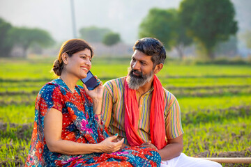 Indian rural woman sitting with husband at agriculture field and talking on smartphone.