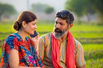 Indian rural woman sitting with husband at agriculture field and talking on smartphone.