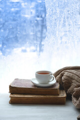 tea cup, book, sweater and winter frozen window abstract background. cozy mood, home comfort in snowy cold weather. festive winter season. Christmas, New Year holidays