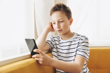 Teenage boy sitting on couch using smartphone texting and having video chat on internet online at home. Child holding mobile phone and looking at screen. Social media. Young blogger influencer