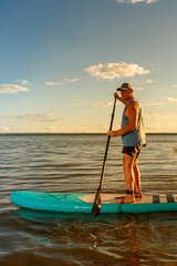 a man in shorts and a T-shirt on a SUP board with an oar floats on the water against the background of the setting sun.