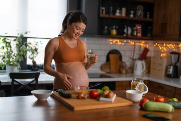 Obraz na płótnie Canvas Pregnant woman holding a glass of water while standing in the kitchen and preparing breakfast at home. Beautiful healthy pregnant woman following healthy lifestyle, caring about health.