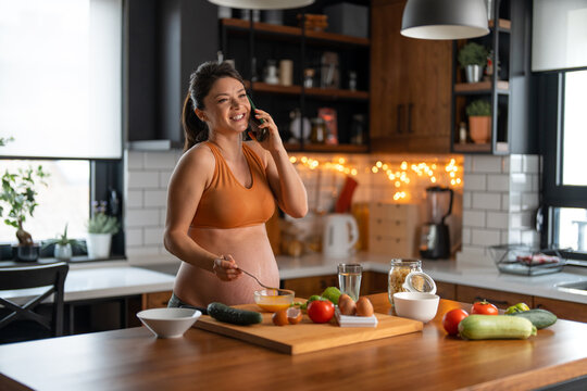 Pregnant woman having a call while making breakfast in the kitchen. Smiling, starting the day with good news.