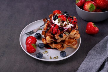 Stack of waffles with strawberry, blueberry, whipped cream and chocolate sauce on ceramic plate on dark gray table. Appetizing dessert