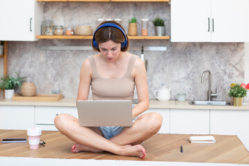 Young beautiful woman in headphones studying online at home. She sits on the table in the kitchen