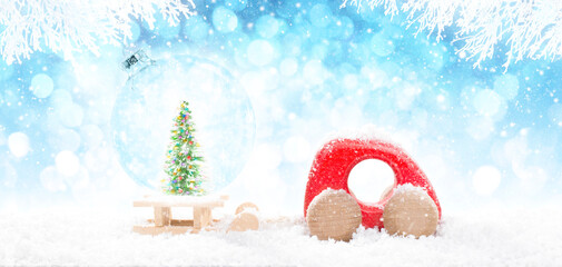 Red Wooden Car Pulling Sled with A Transparent Christmas Ball with green colorful glitter pine Christmas tree in perspective on bright blue bokeh winter background with white branches and falling snow