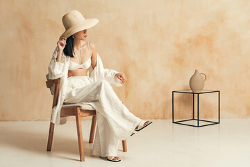Unrecognizable asian female in big hat and beach suit sitting on chair in minimalistic decor