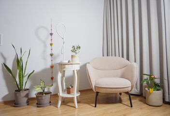 Elegante mueble de lectura beige, acompañado de maceteas con plantas y mesa blanca con lampara y adornos con fondo blanco y cortinas grises - obrazy, fototapety, plakaty
