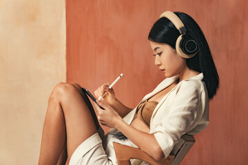 Profile view of asian woman in headphones drawing on tablet with stylus pen, sitting on chair