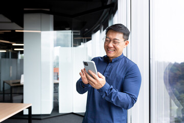 Naklejka na ściany i meble Asian businessman inside the office in a casual shirt and glasses is using the phone, the boss is holding a smartphone in his hands reading news and typing a message smiling.