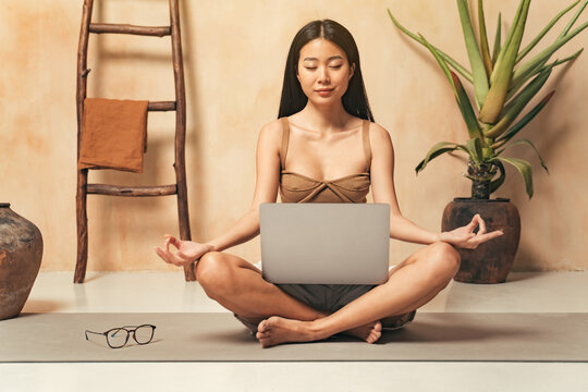 Asian girl sitting in lotus pose with laptop, watching yoga tutorial online, using meditation app