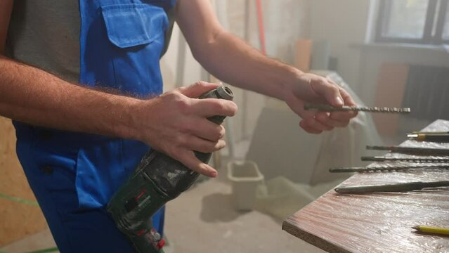 Man in blue construction overalls puts spiral borer on perforator. Builder installing drill boer and checking its work against backdrop of room with building materials and tools. Close up.