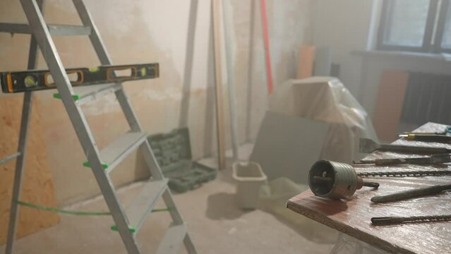 Room under renovation. Ladder with water level, wooden table with metal nozzles for perforator or electric drill. Spiral borers for drilling and bowl for cutting holes for socket.