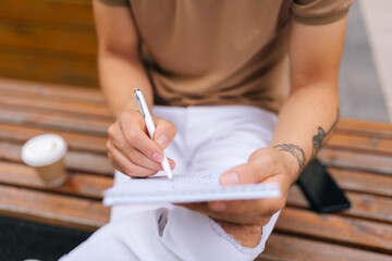 High-angle cropped shot of stylish male artist with tattooed hands sitting on bench and making sketches with pen on paper. Talented young man with pencil and sketchbook drawing outdoors. - Powered by Adobe