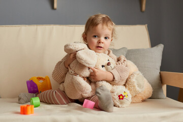 Horizontal shot of cute blonde baby girl in beige jumper sitting on sofa with a plush big toys, looking at camera, playing alone at home, posing among multicolored sorter parts.