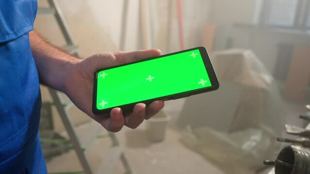 Male builder holding touch screen smartphone with green screen chroma key. Man using finger for touching, clicking, swiping, sliding, zooming, dragging on phone screen. Close up.