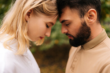 Fototapeta premium Close-up of loving married couple standing foreheads touched with closed eyes. Happy young bearded man and pretty blonde woman on romantic date in summer park. Couple relaxing heads embracing.