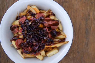 Top view of fried potatoes with sausages, red cabbage, cheddar cheese and barbecue sauce, in a white bowl with a wooden background.
