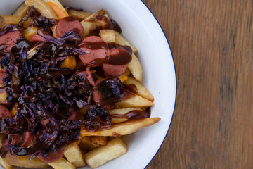 Top view of fried potatoes with sausages, red cabbage, cheddar cheese and barbecue sauce, in a white bowl with a wooden background.