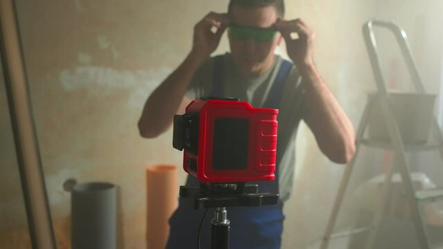 Man in blue construction overalls and goggles adjusts red laser roulette with flashing green beam. Intersecting green beam of laser tape measure is displayed on brown wall.