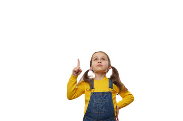 Caucasian lovely little girl with two ponytails, in bright yellow sweater and blue denim overalls, looks up and points forefinger at copy space on white background, for advertising promotional text