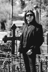 Stylish dark gothic guy with black clothes in front of a grave in cemetery in a sunny day (in black and white)