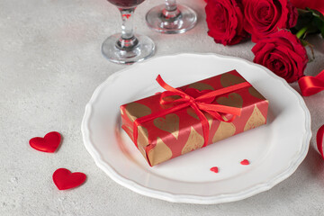 Gift in wrapping paper on white plate, red roses and two glasses wine, concept for Valentines Day